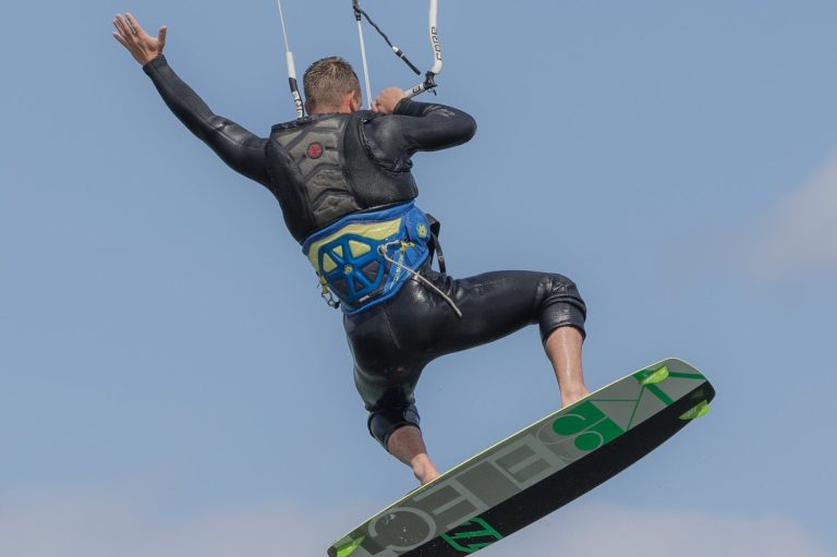 Kitesurfer springt mit Brett über das Wasser, blauer Himmel im Hintergrund.